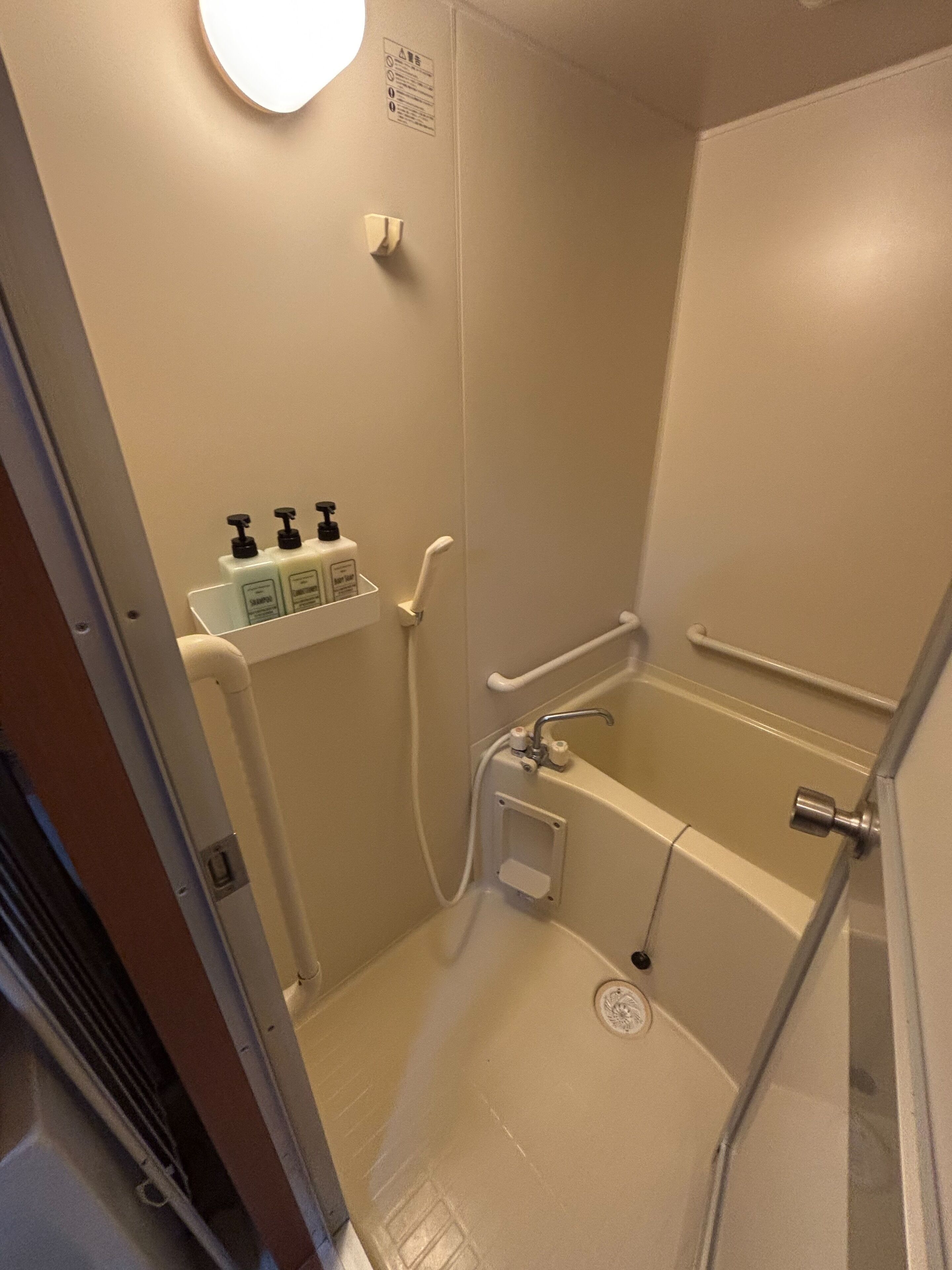 A small, enclosed shower area with a white bathtub, a handrail, and a shelf with toiletries.