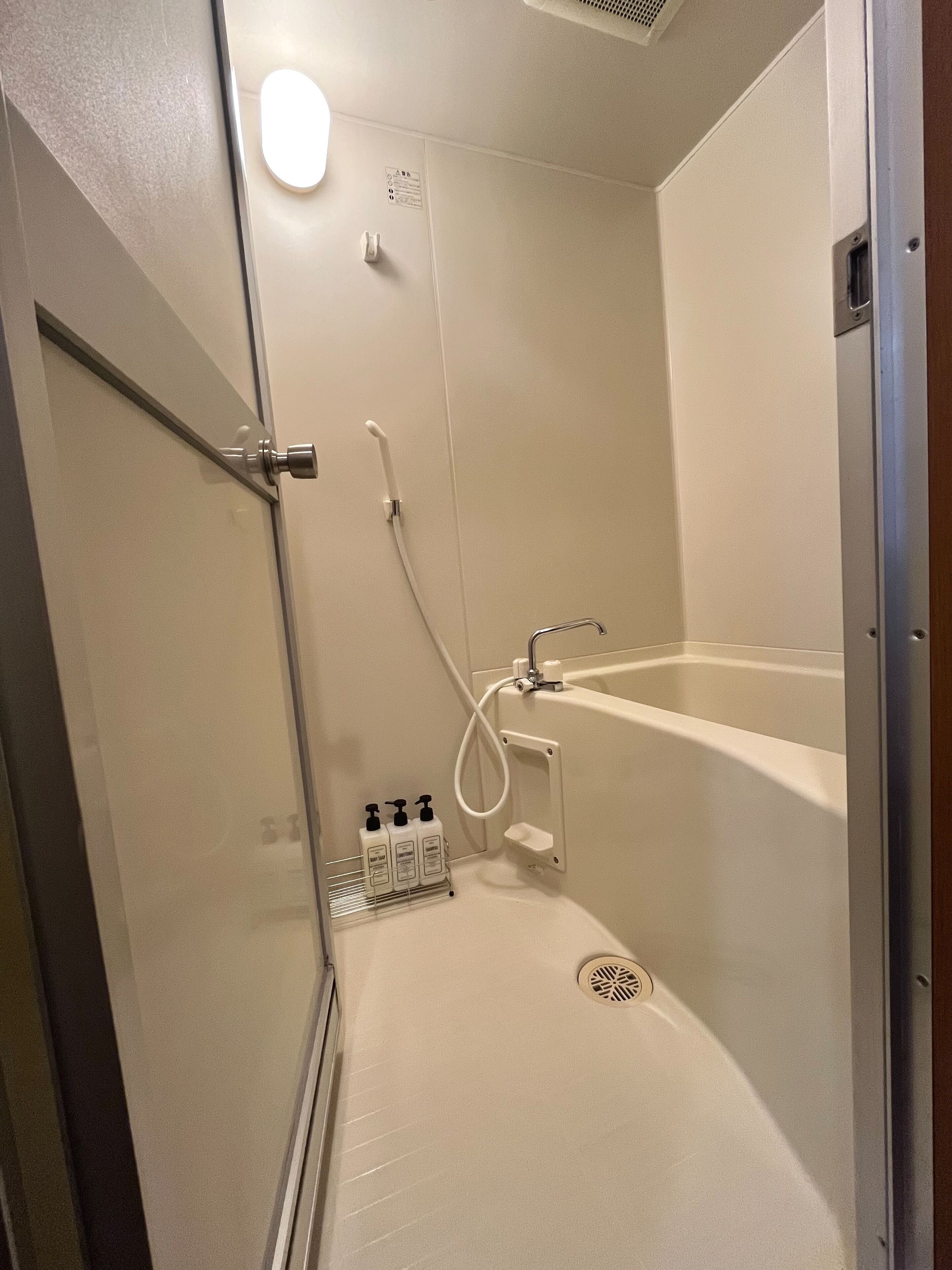 A walk-in shower with a glass door, a handheld showerhead, a handheld faucet, and a small shelf with toiletries.