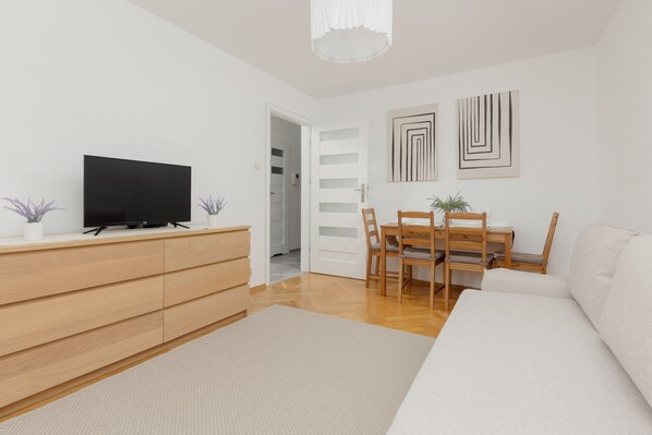 Een moderne woonkamer met een bank, televisie, houten eettafel, stoelen en een witte muur met abstracte kunst.