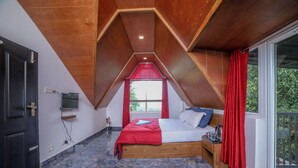 Een slaapkamer met een schuine kap, een bed met een rode deken, een houten nachtkastje en een raam met rode gordijnen.
