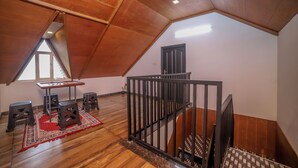 Een zolderkamer met een schuine kap, een houten vloer, een kleine tafel met krukjes, een rood, geruit tapijt en een zwarte leuning.