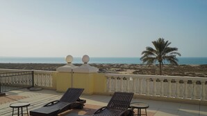 Un balcon avec deux fauteuils et une petite table, donnant sur une plage bordée d’un palmier et sur la mer.