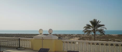 A balcony with two lounge chairs and a small table, overlooking a beach with a palm tree and the sea.
