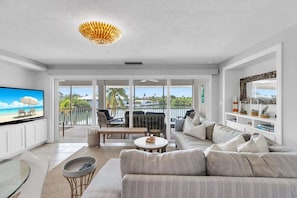 A spacious living room with a large flat-screen TV, a sofa, a coffee table, and a balcony with a view of the beach.