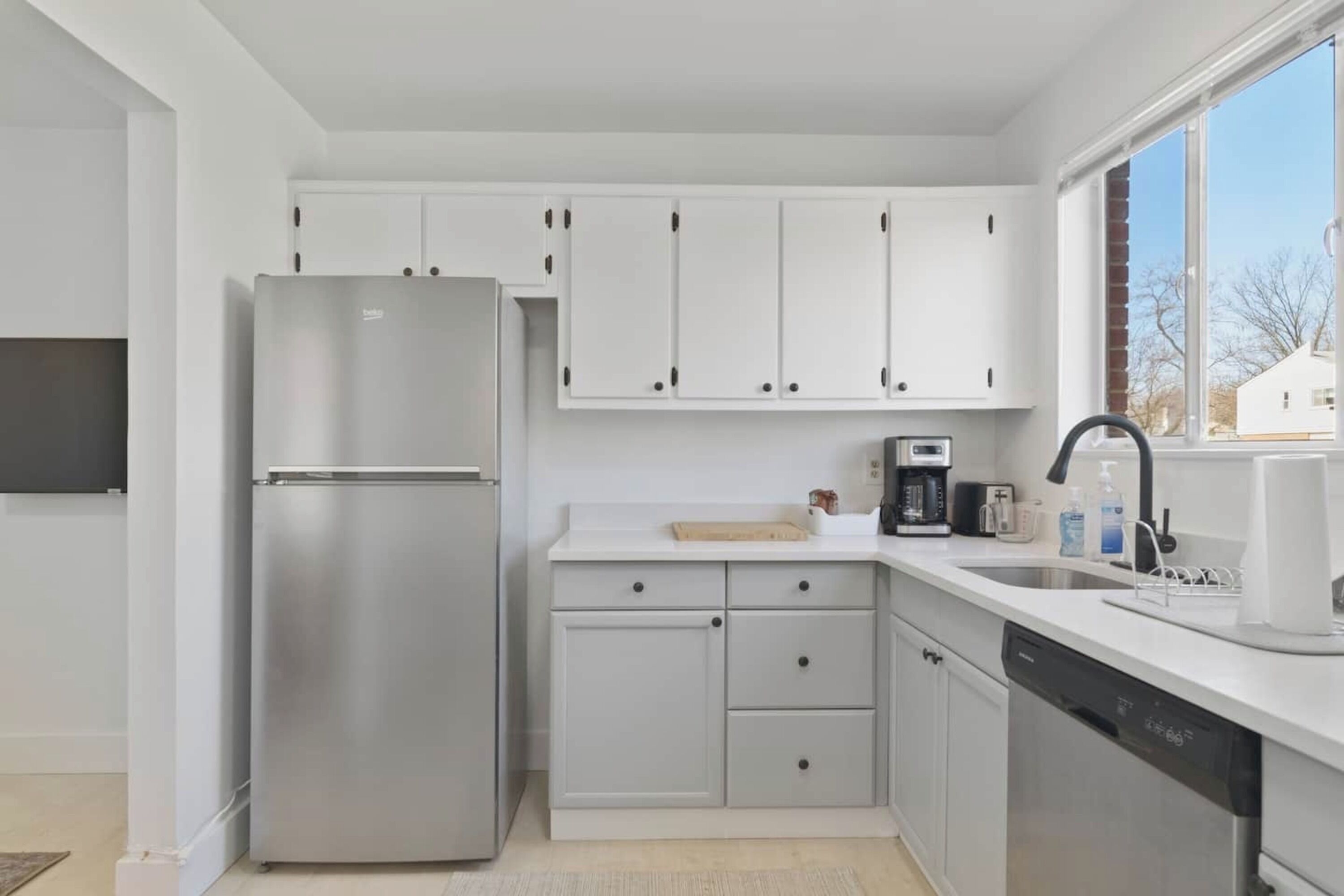 A modern kitchen with white cabinets, a stainless steel refrigerator, a dishwasher, and a window with a view of houses.