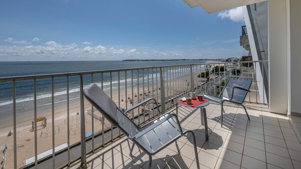 Ein Balkon mit Blick auf Strand und Meer. Zwei Stühle und ein kleiner Tisch mit einer roten Matte sind vorhanden.