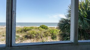 Vista a través de una ventana de una playa con cielo despejado, orilla arenosa y algo de vegetación.