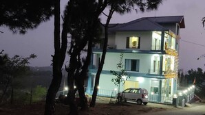 Un bâtiment à deux étages avec un balcon à l’étage supérieur, une voiture garée devant et des arbres au premier plan.