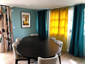 A dining area with a round table and six chairs, teal curtains, and a framed picture on the wall. - Gite LE Passiflore (Beuvron-en-Auge)
