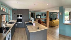 A modern kitchen with a central island, a dining area with a yellow sofa, and a large window offering a view of the outdoors. - Gite LE Passiflore (Beuvron-en-Auge)