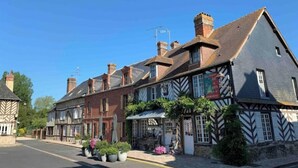 A traditional half-timbered building with a pub on the ground floor, a row of potted plants, and a clear blue sky. - Gite LE Passiflore (Beuvron-en-Auge)