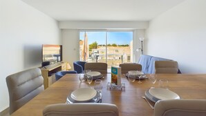 A dining area with a large table set for four, a television on the left, and a view of buildings through the window. - Flat for 4 in the town centre - near by tren station (Saint-Hilaire-de-Riez)