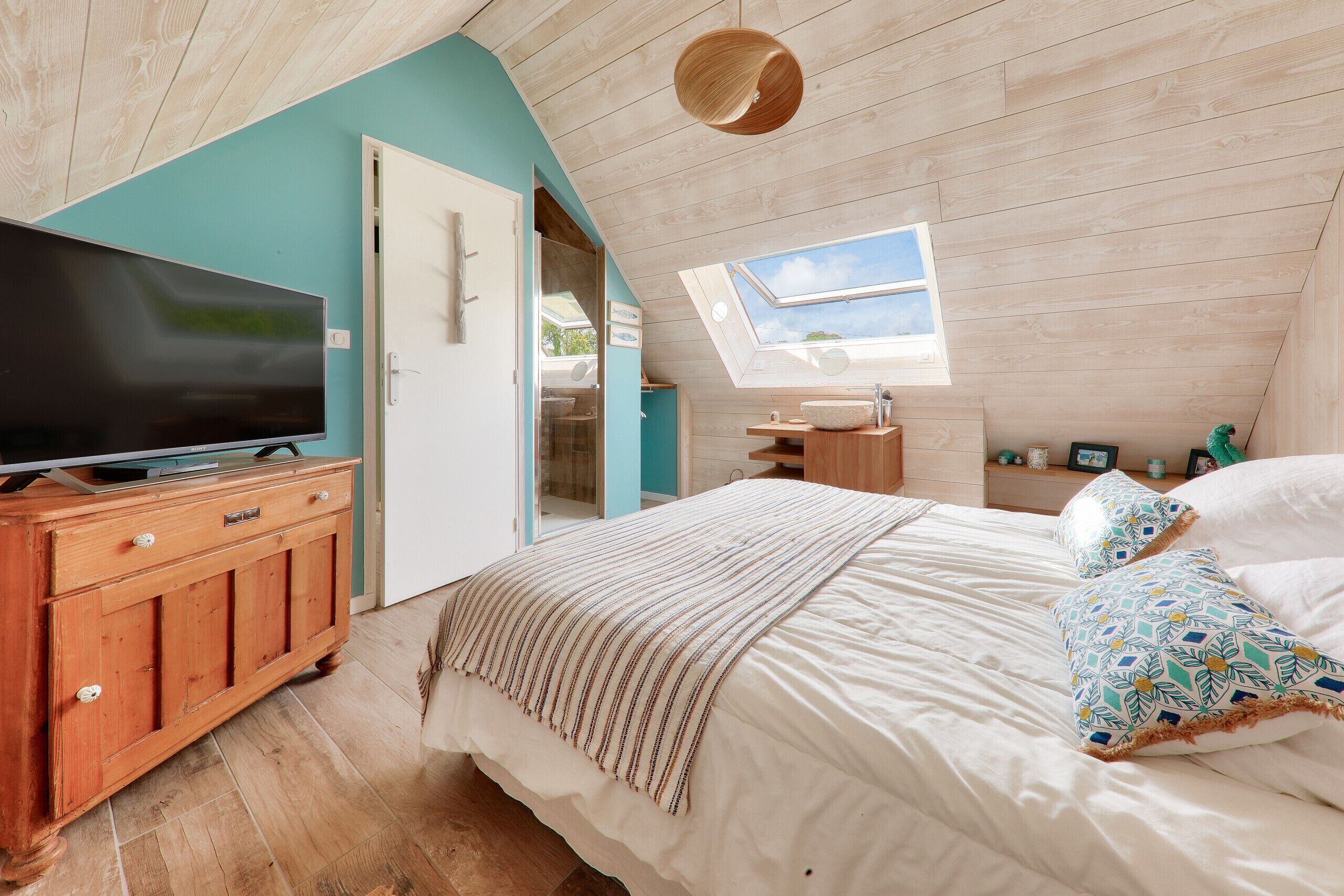 Une chambre avec une télévision, une armoire en bois, un lit agrémenté d’oreillers à motifs et une lucarne.