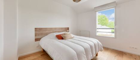 A bedroom with a wooden floor, a bed with white bedding, a woven light fixture, and a window with a view of greenery.