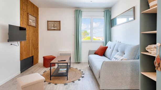 Un salon moderne avec un canapé gris, une petite table, un pouf rouge, un téléviseur fixé à un mur en bois et une fenêtre avec des rideaux.
