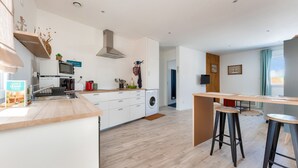 Une cuisine moderne équipée d’un four à micro-ondes, d’un four et d’un évier. On y trouve une table à manger en bois avec deux tabourets. La pièce est dotée d’un parquet en bois clair et de murs blancs.