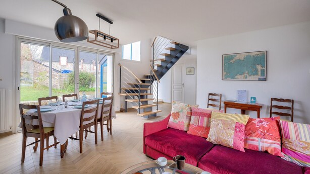 A spacious living area with a dining table set for four, a red sofa with multiple cushions, a staircase, and a large window offering a view of the outdoors. - Quiet house for 6 - Less than 2km from the beach (Dinard)