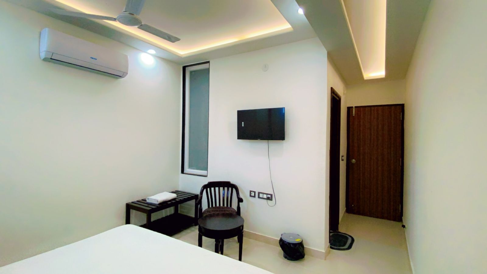 A hotel room with a bed, a desk with a chair, a TV mounted on the wall, an air conditioner, and a door.