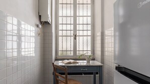 Una cocina con paredes de azulejos blancos, una ventana con vidrio esmerilado, un refrigerador blanco, una silla de madera y una pequeña mesa con un libro y una planta en maceta.