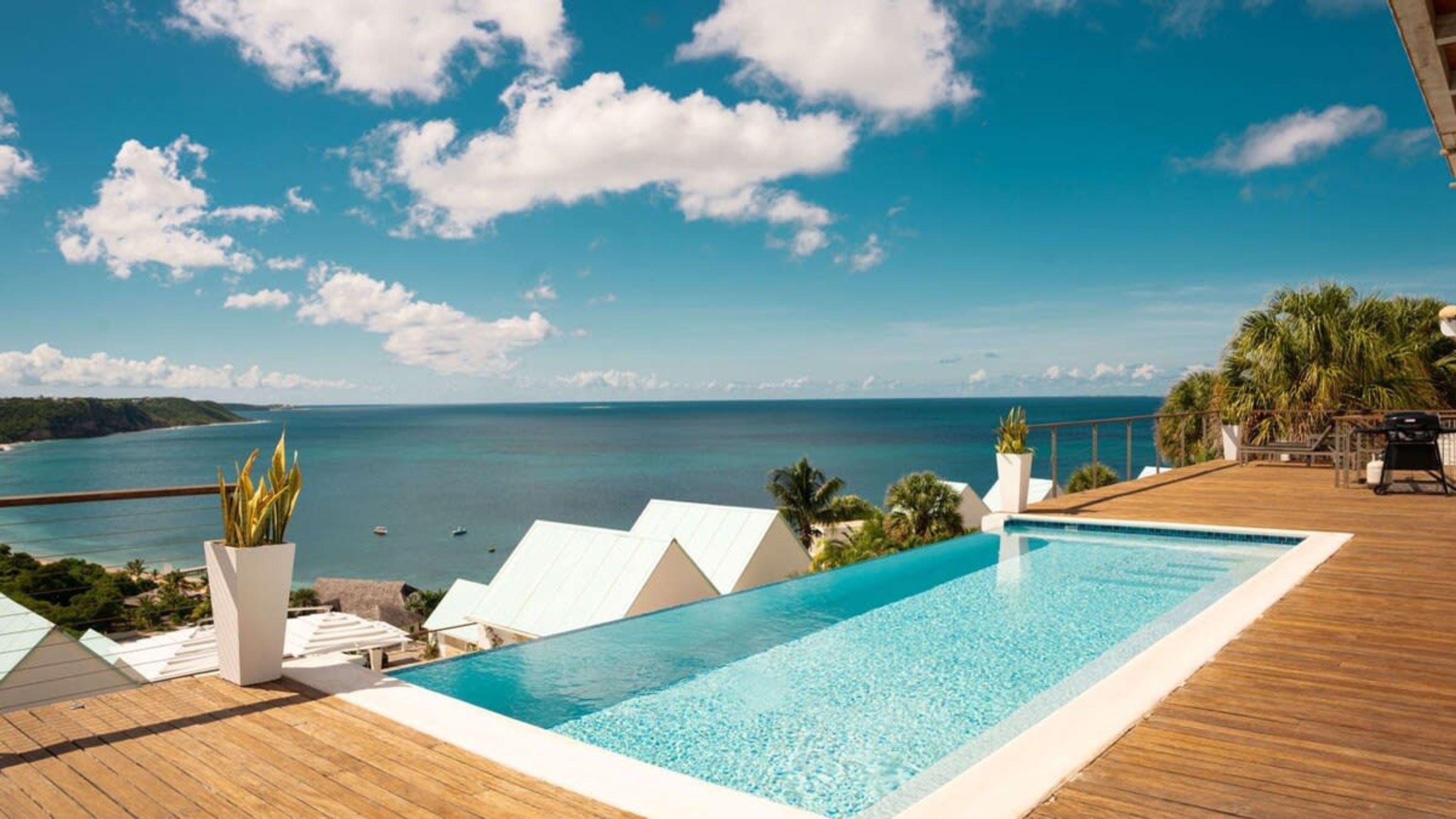 A pool with a view of the sea, wooden deck, lounge chairs, and a white building with a triangular roof.
