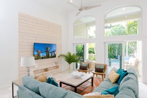 A spacious living room with a large sofa, a coffee table, and a TV mounted on the wall. There is a large window providing a view of the outdoors. - Waterfront Oasis W Boat Dock Heated Pool (Key Colony Beach)