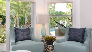 Ein Zimmer mit zwei hellblauen Sesseln, einem Beistelltisch mit Lampe und Pflanze, und Blick auf einen Pool und Palmen durch große Fenster.