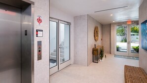 Ein Innenraum mit Aufzug, Glastüren, einer Treppe, einer Rezeption und Blick ins Freie.