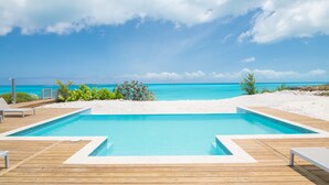 Una piscina con cubierta de madera, rodeada de arena blanca y vegetación, con vistas a una playa y al océano.