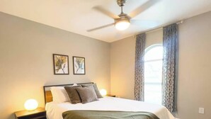 Une chambre à coucher comprenant un lit, deux tables de chevet, un ventilateur de plafond et une fenêtre avec des rideaux.