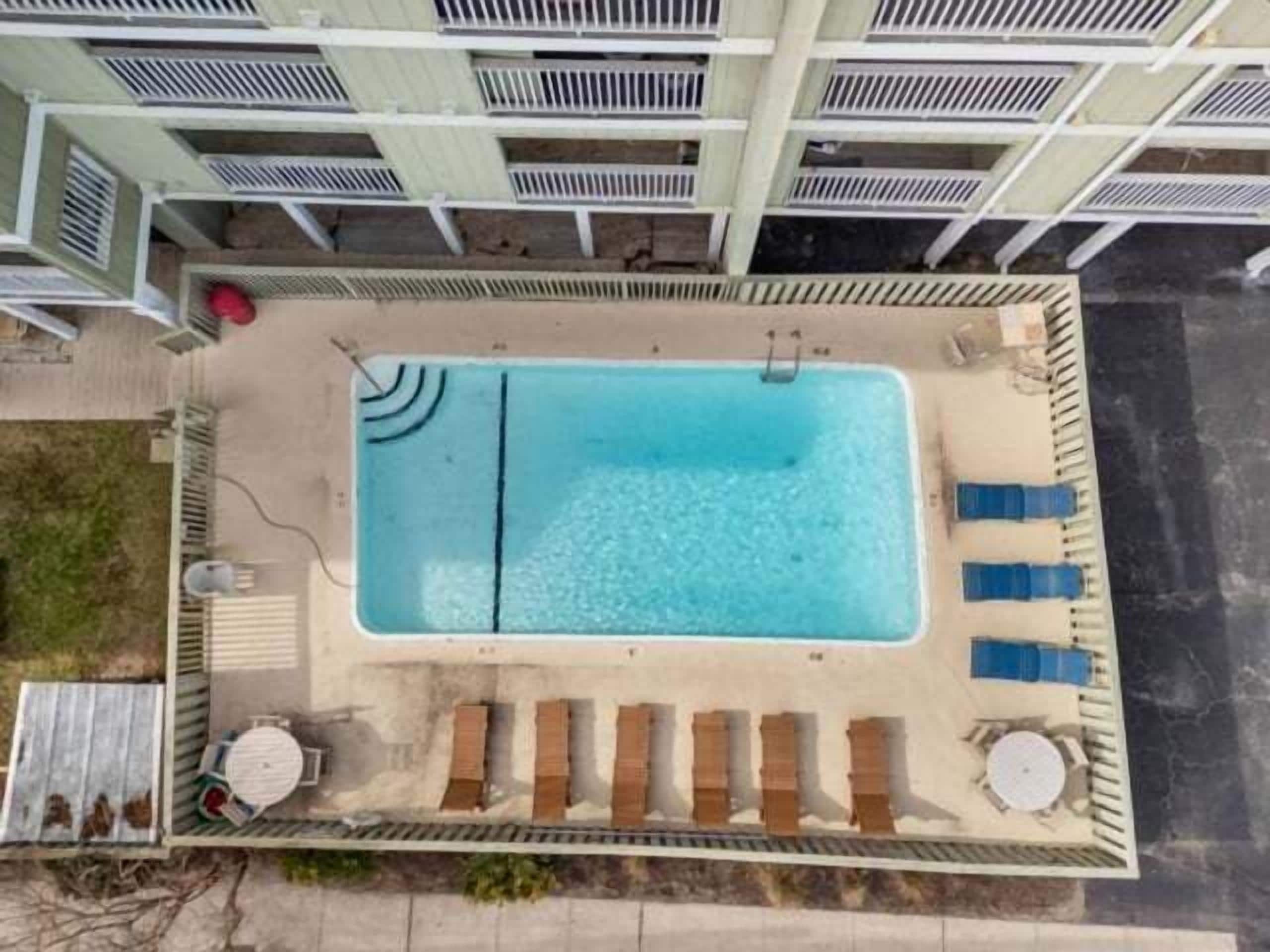 Oversigtsbillede af en swimmingpool med liggestole og solstol, omgivet af et hegn med en bygning i baggrunden.