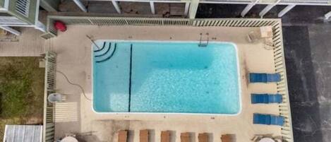 Oversigtsbillede af en swimmingpool med liggestole og solstol, omgivet af et hegn med en bygning i baggrunden.