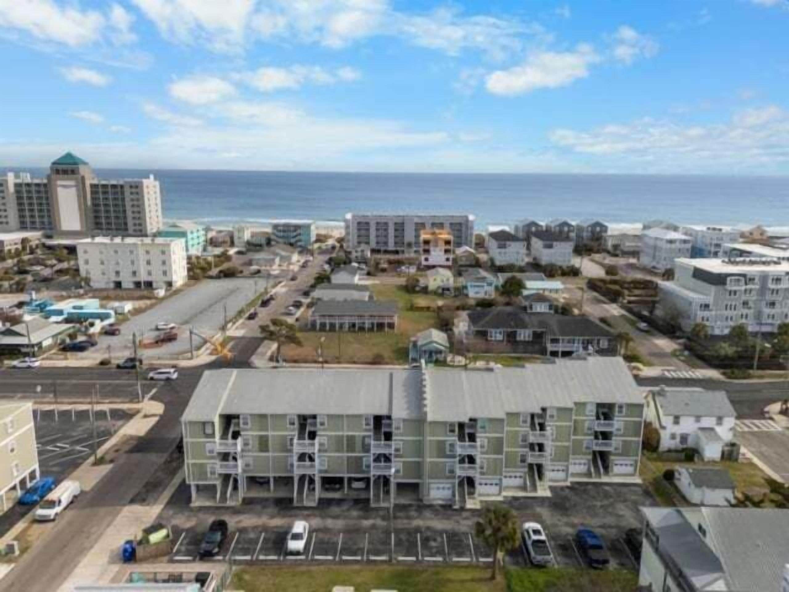 A coastal hotel with multiple-story buildings, a parking lot, and a view of the ocean.