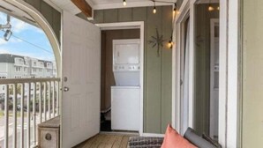 A balcony with a wooden floor, a sofa with a cushion, a small table, and a string of lights. - Sea The Good 2 Bedroom Condo by RedAwning (Carolina Beach)