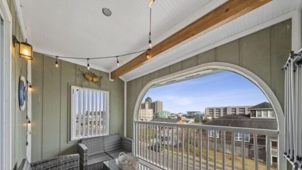 A covered balcony with wicker furniture, a glass coffee table, and a view of buildings in the distance. - Sea The Good 2 Bedroom Condo by RedAwning (Carolina Beach)