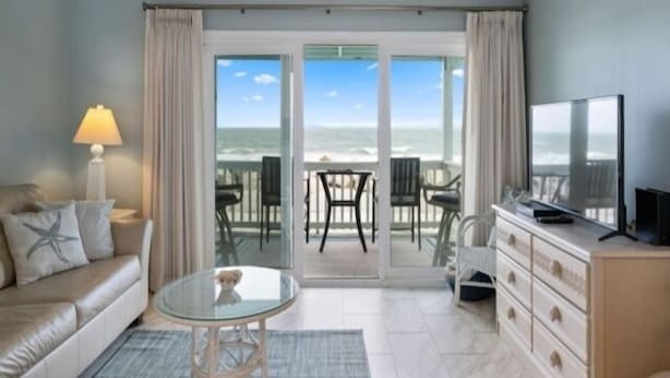 A room with a glass door leading to a balcony with a view of the ocean. There is a sofa, a small round table, and a TV on a cabinet. - Beach Bliss 1 Bedroom Condo by RedAwning (Carolina Beach)