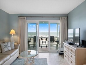 A room with a glass door leading to a balcony with a view of the ocean. There is a sofa, a small round table, and a TV on a cabinet. - Beach Bliss 1 Bedroom Condo by RedAwning (Carolina Beach)