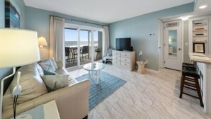 A modern living room with a sofa, a coffee table, a TV, and a balcony with a view of the sea. - Beach Bliss 1 Bedroom Condo by RedAwning (Carolina Beach)