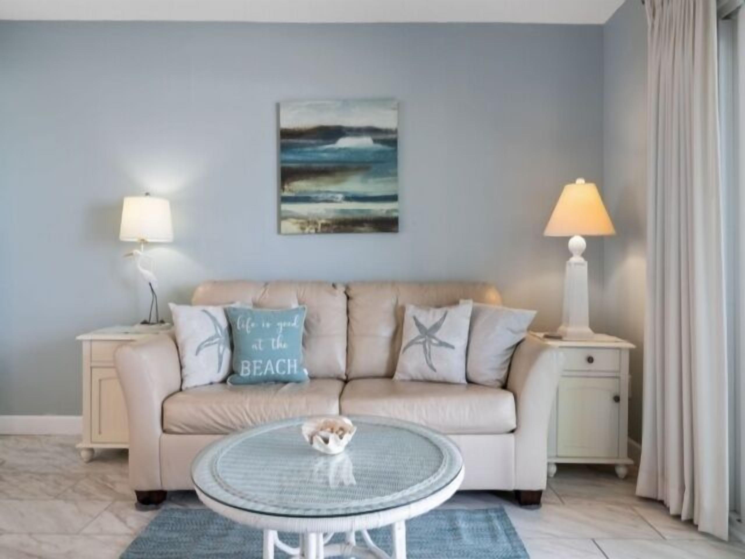 A living room with a beige sofa, two side tables with lamps, a glass coffee table, and a beach-themed pillow.