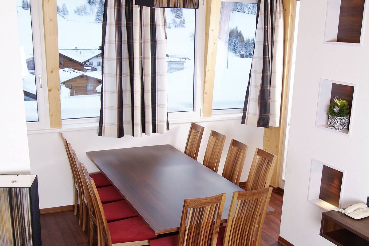 A dining area with a long wooden table, red cushioned chairs, and a view of a snowy landscape through large windows.