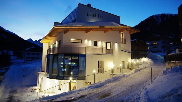 A modern building with a sign 'astellina' on the roof, surrounded by snow and mountains. - Astellina hotel-apart (Ischgl)