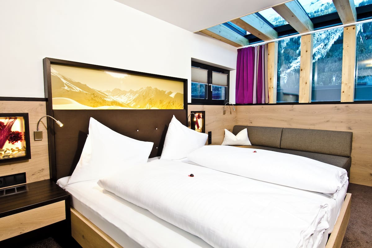 A modern hotel room with a large bed, a headboard with a mountain view, a skylight, and a window with a view of snow-capped mountains.