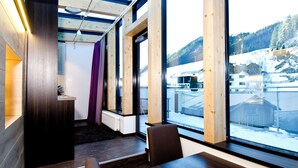 A modern interior with a wooden desk, a chair, and a large window offering a view of a snowy landscape and buildings. - Astellina hotel-apart (Ischgl)