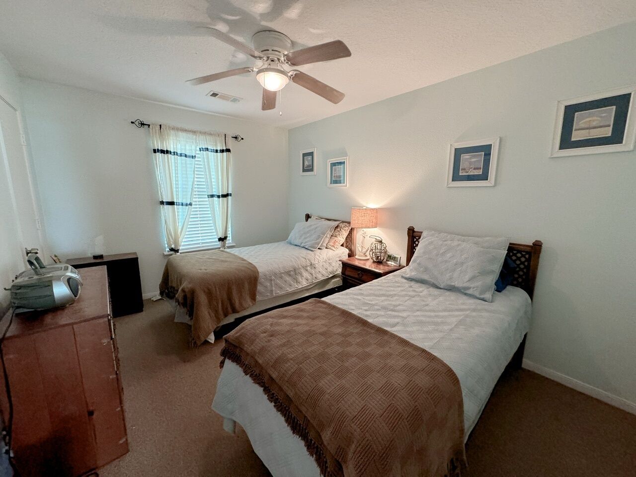 Une chambre avec deux lits simples, un ventilateur de plafond, un bureau avec une lampe et une fenêtre avec des rideaux.