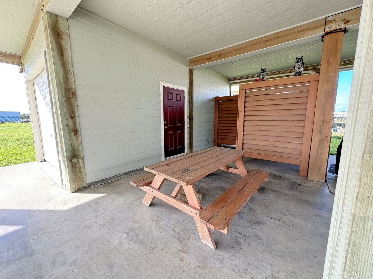 Una mesa de picnic de madera en un espacio exterior cubierto, con una puerta roja y una valla de madera.