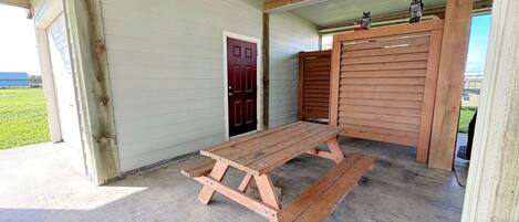 Una mesa de picnic de madera en un espacio exterior cubierto, con una puerta roja y una valla de madera.
