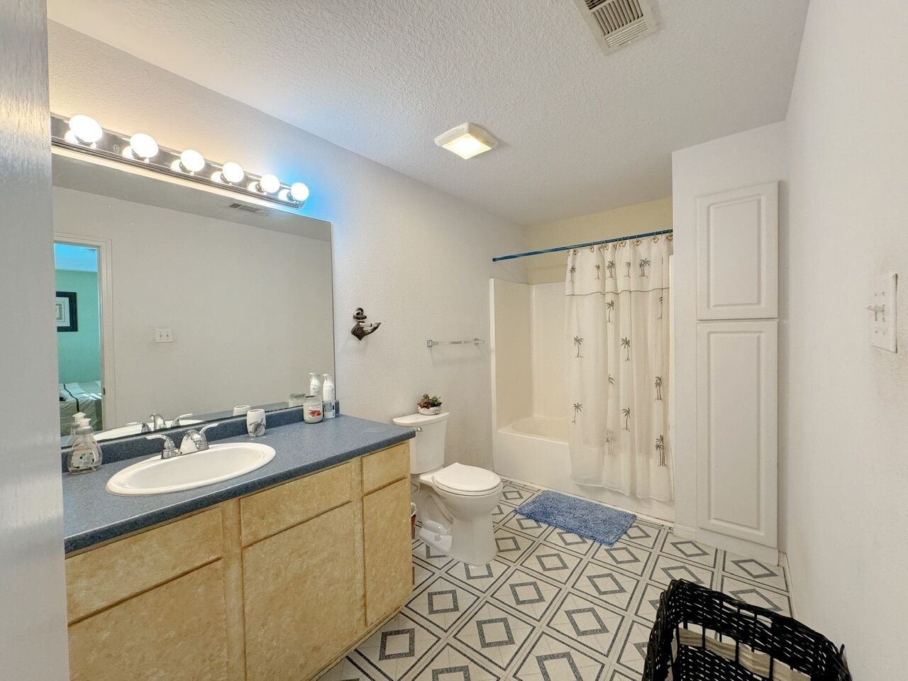 Un baño con lavamanos, inodoro y ducha, con piso de baldosas geométricas y un tapete azul.