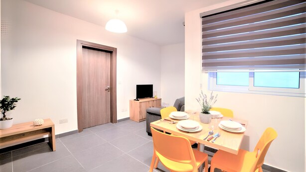 A modern dining area with a wooden table set for four, surrounded by orange chairs. A television is placed on a wooden cabinet against the wall. - Standard Apartment in Gzira N33 (Il-GÅ¼ira)
