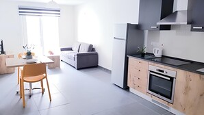 A modern kitchen with a built-in oven, a refrigerator, and a coffee maker. There is a dining area with a table and chairs, and a gray sofa in the background. - Comfort Apartment in Gzira N112 (Il-GÅ¼ira)
