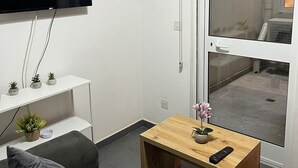 Una sala de estar moderna con un televisor de pantalla plana, una mesa de centro de madera con un control remoto, un sofá gris y un ventanal corredizo que da a un balcón.