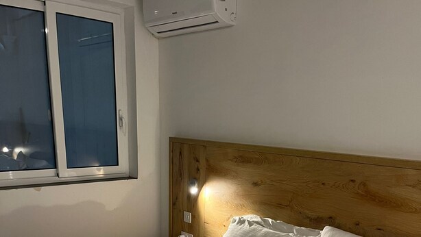 Una cama bien hecha con sábanas y almohadas blancas, un cabecero de madera, una ventana con persianas y un equipo de aire acondicionado en la pared.